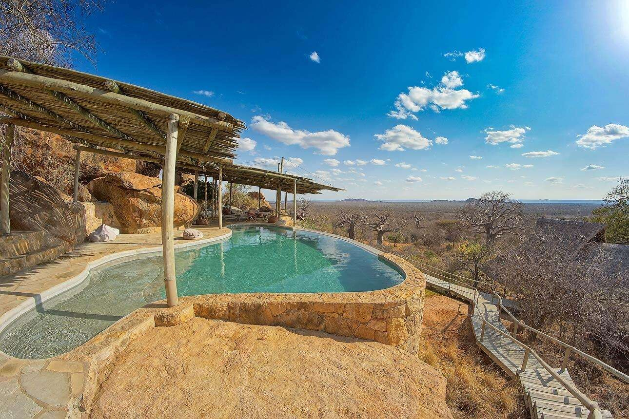 Stone-edged pool on a rocky terrace with a wooden pergola, overlooking a vast dry savannah under a bright blue sky.