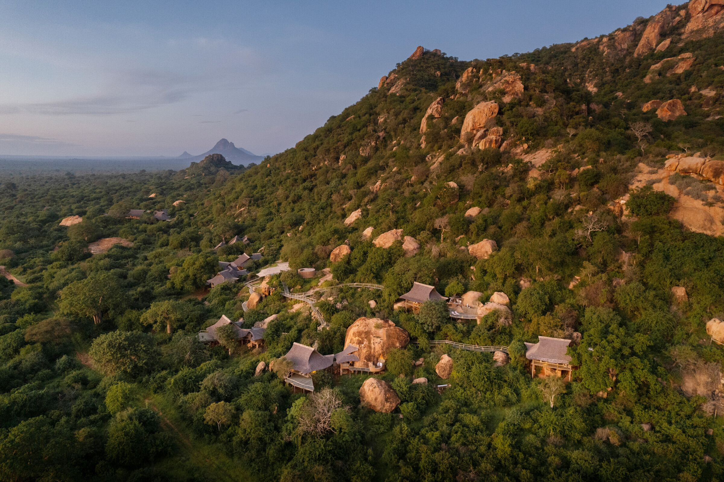 Aerial view of a forested hillside with large reddish boulders, and a cluster of cabins with gray roofs at the base along a winding path.